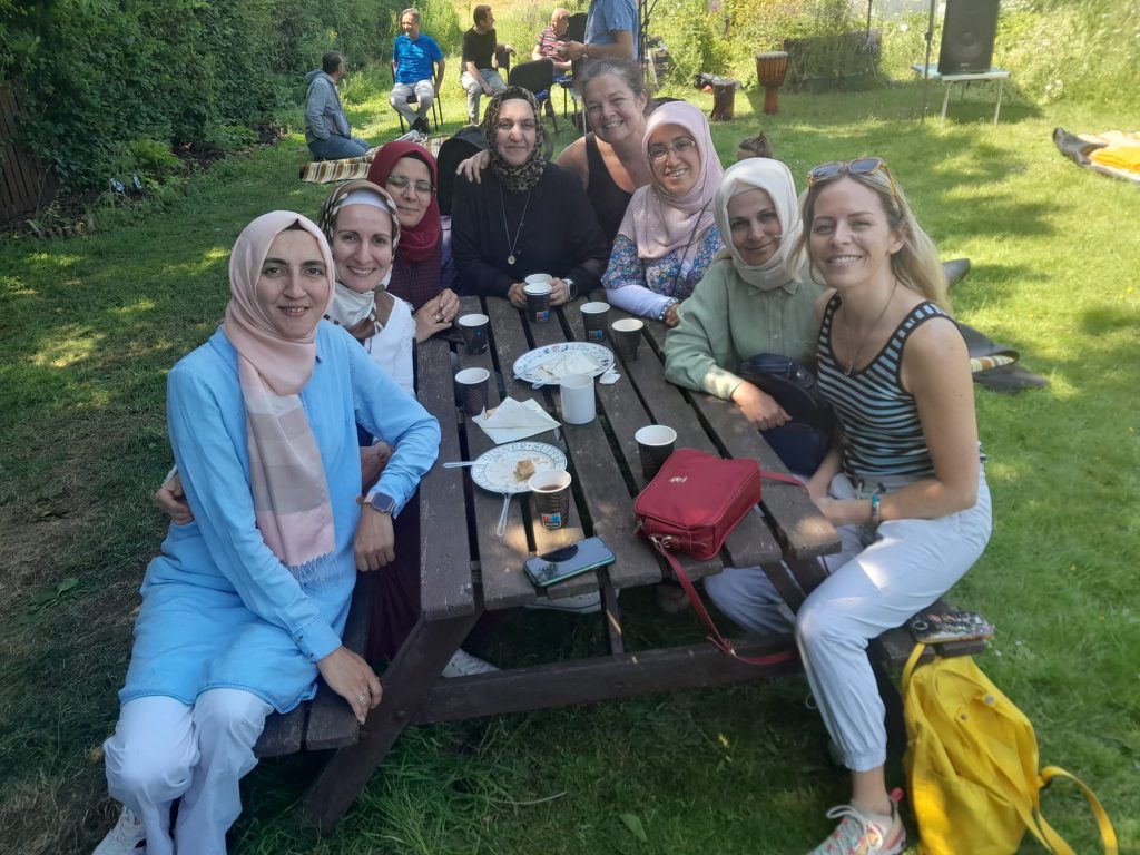 Eight women sat on benches at a table. They're outside. Six of the women are wearing hijabs, the other two have their hair uncovered. All the women are smiling and looking at the camera