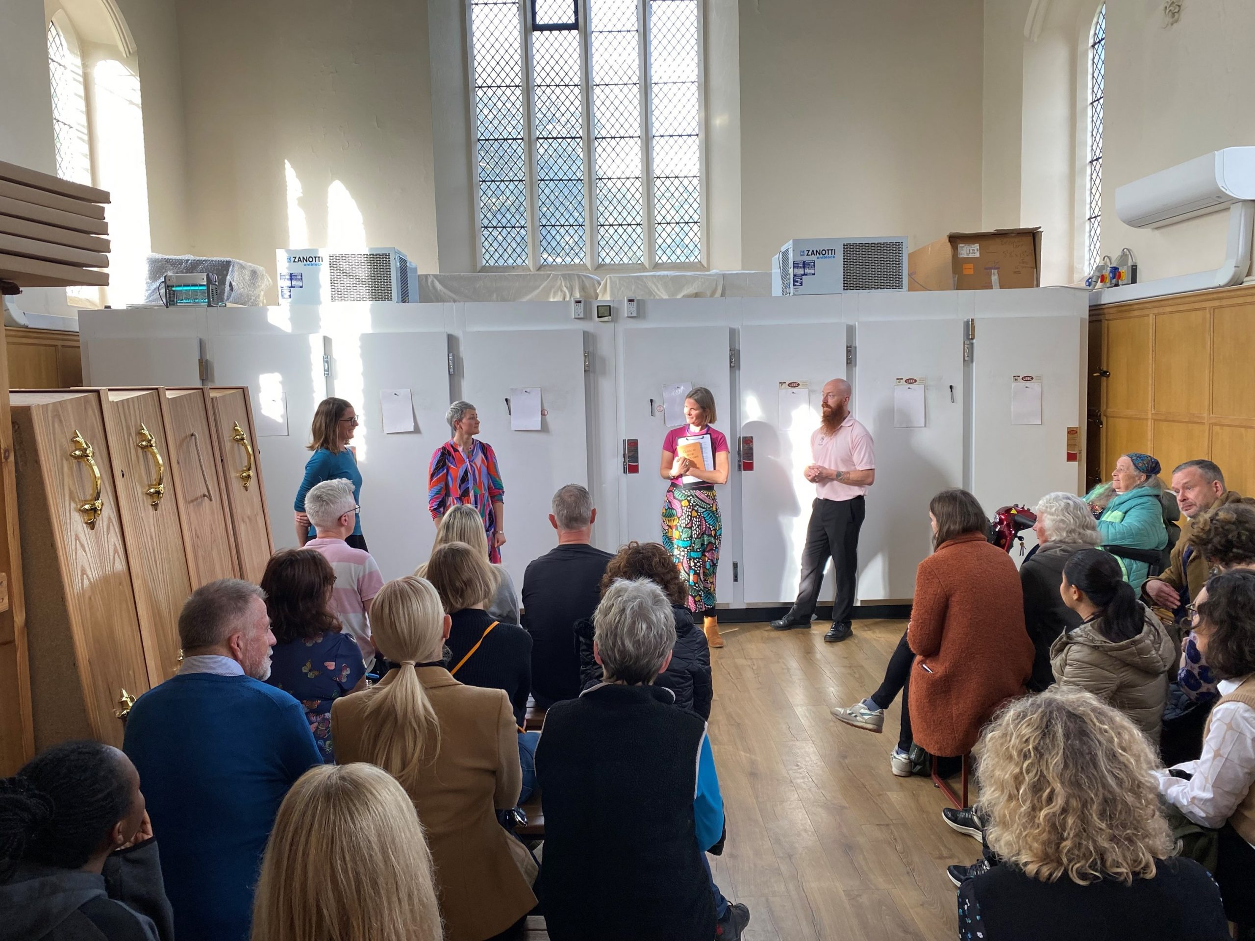 People gathered in a church with coffins to one side. Four people are stood presenting at the front whilst others are gathered on seats, listening
