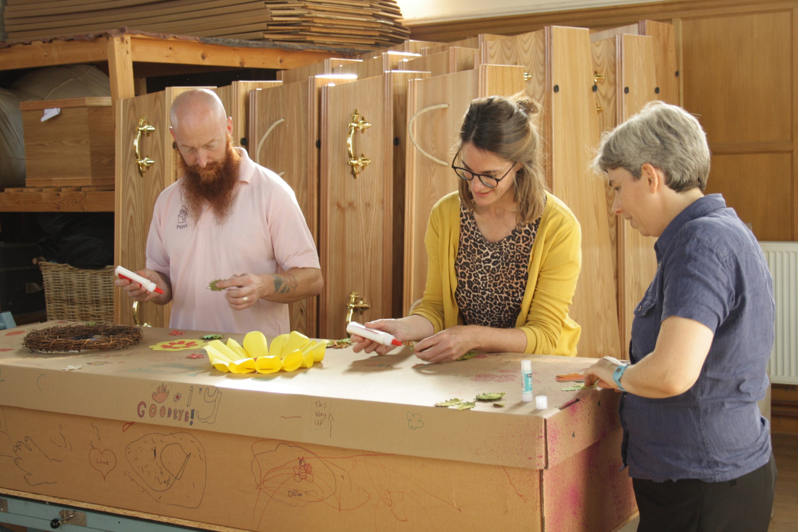 Three people decorating a cardboard coffin using glue, pens and wraths. There are children's drawings on the coffin and a message that reads:'Ivy, goodbye!'