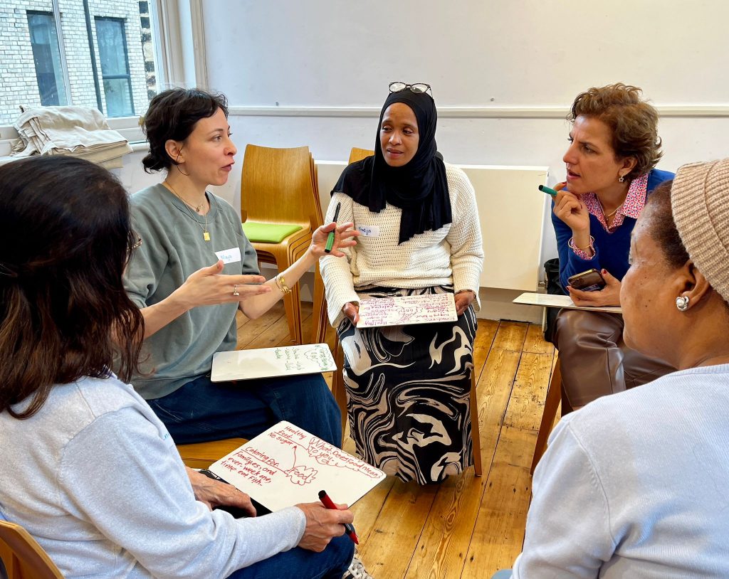 A group of women of different ethnicities sat in a circle on chairs talking with one another. Each woman has a whiteboard on her lap on which she has written 